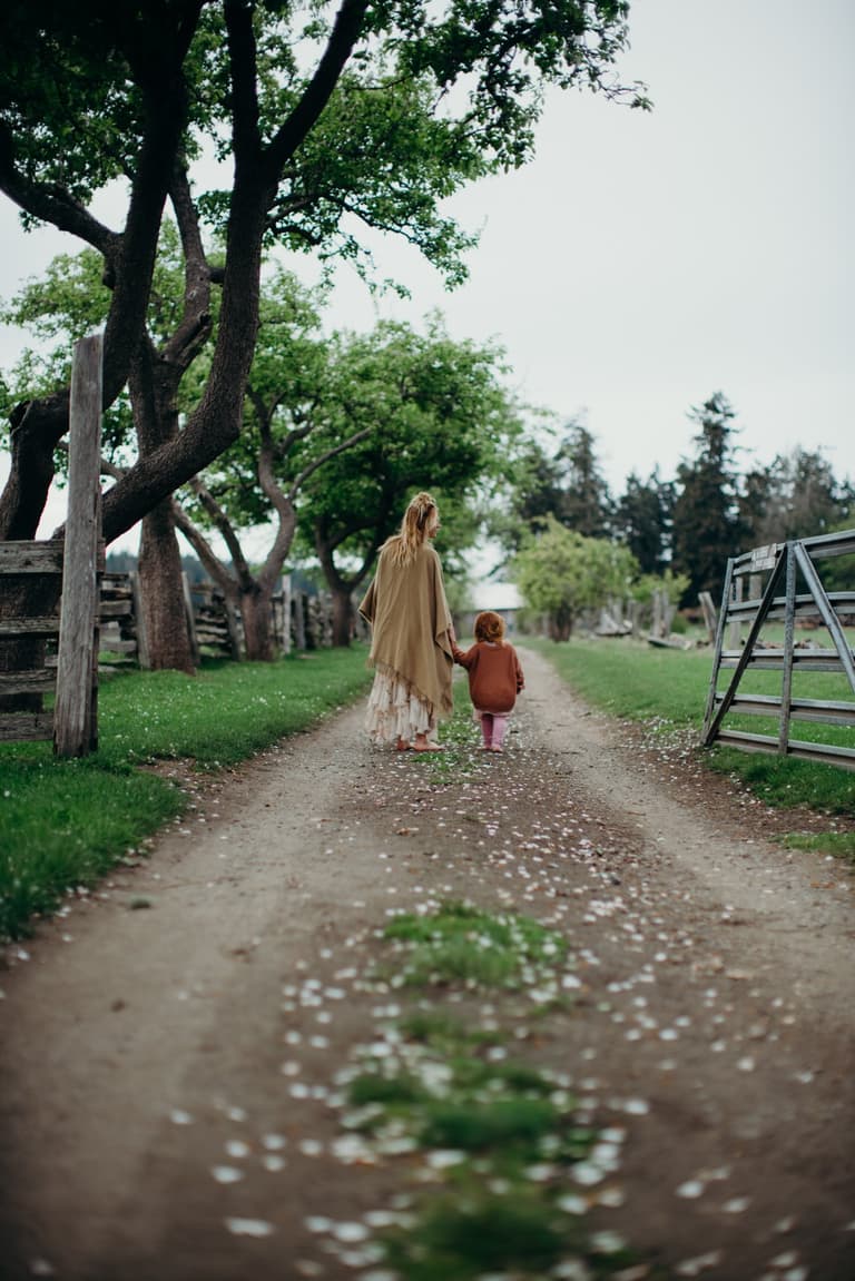 Woman and toddler holding hands on a tree-lined gravel path, soft overcast light filtering through green canopy on Salt Spring Island Farm